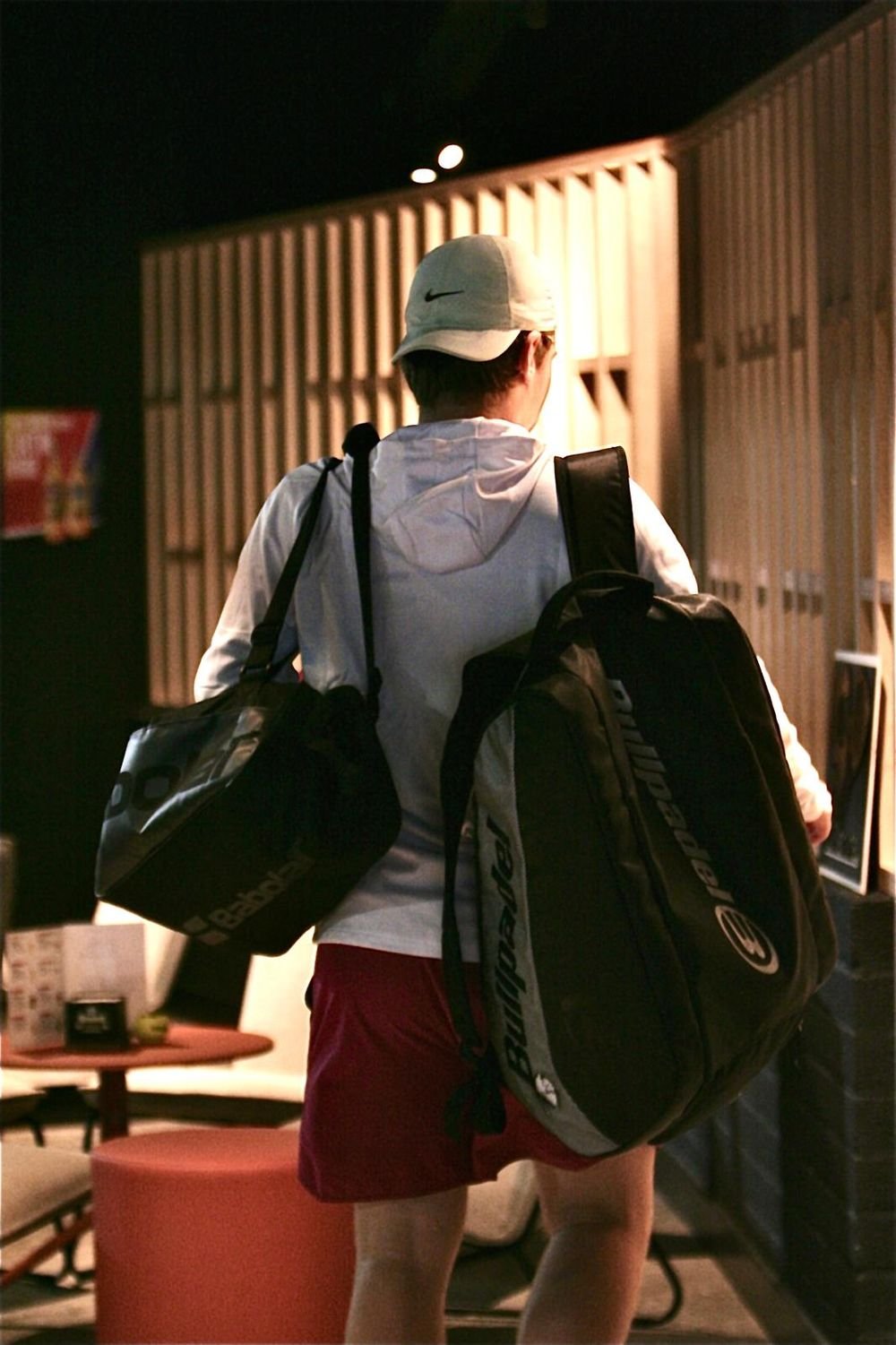Matteo walking into the club from behind, a Babolat holdall on one shoulder and a Bullpadel racket bag on the other